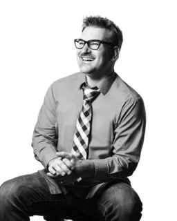 black and white image of a man smiling clasping hands while seated wearing glasses and a tie